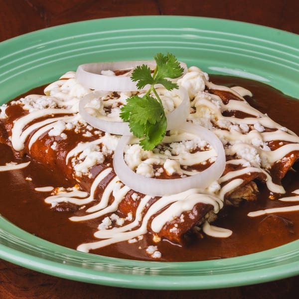Three enchiladas covered in rich mole sauce, crema, crumbled cheese, onion rings, and cilantro.