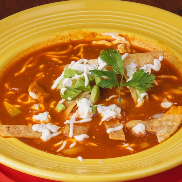 A vibrant yellow bowl of Los Alebrijes' tortilla soup with avocado, cheese, and cilantro.