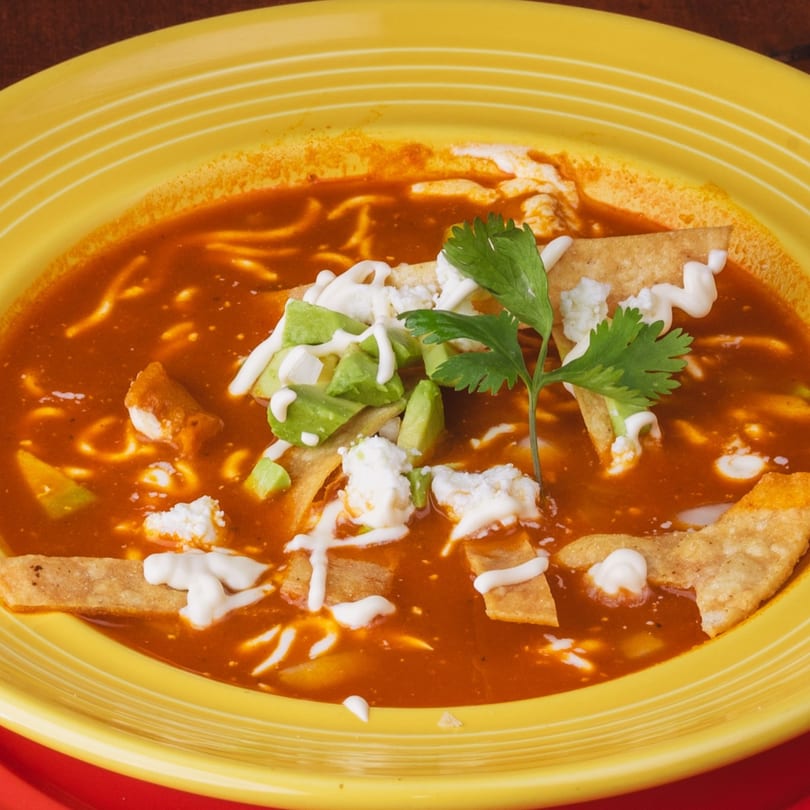 A vibrant yellow bowl of Los Alebrijes' tortilla soup with avocado, cheese, and cilantro.