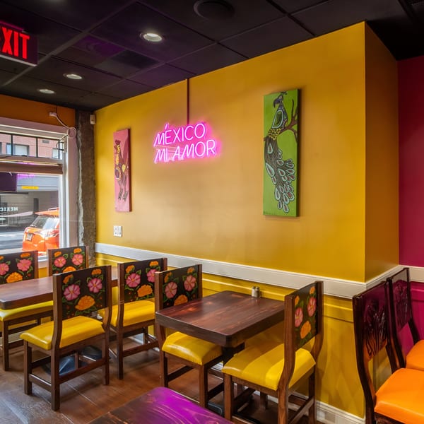 Vibrant Mexican restaurant interior with yellow walls, neon "México Mi Amor" sign, and colorful floral chairs.